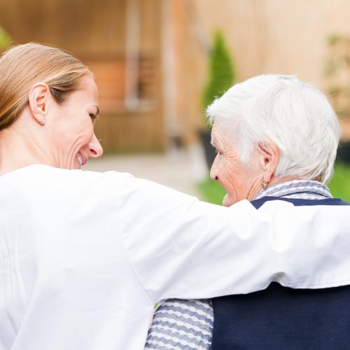 Eine Pflegering legt ihren Arm und eine ältere Frau mit kurzen grauen Haaren und lächelt sie an