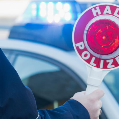 Ein Polizist hält eine rote Stoppkelle hoch. Im Hintergrund sieht man das Blaulicht eines Einsatzfahrzeugs