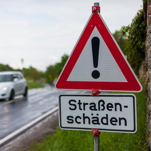 Bild einer kaputten Straße. Im Vordergrund steht ein Warnschild mit der Aufschrift "Straßenschäden"