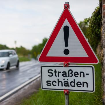Bild einer kaputten Straße. Im Vordergrund steht ein Warnschild mit der Aufschrift "Straßenschäden"