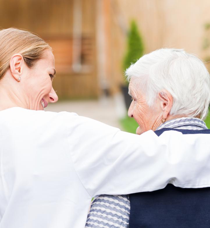 Eine Pflegering legt ihren Arm und eine ältere Frau mit kurzen grauen Haaren und lächelt sie an