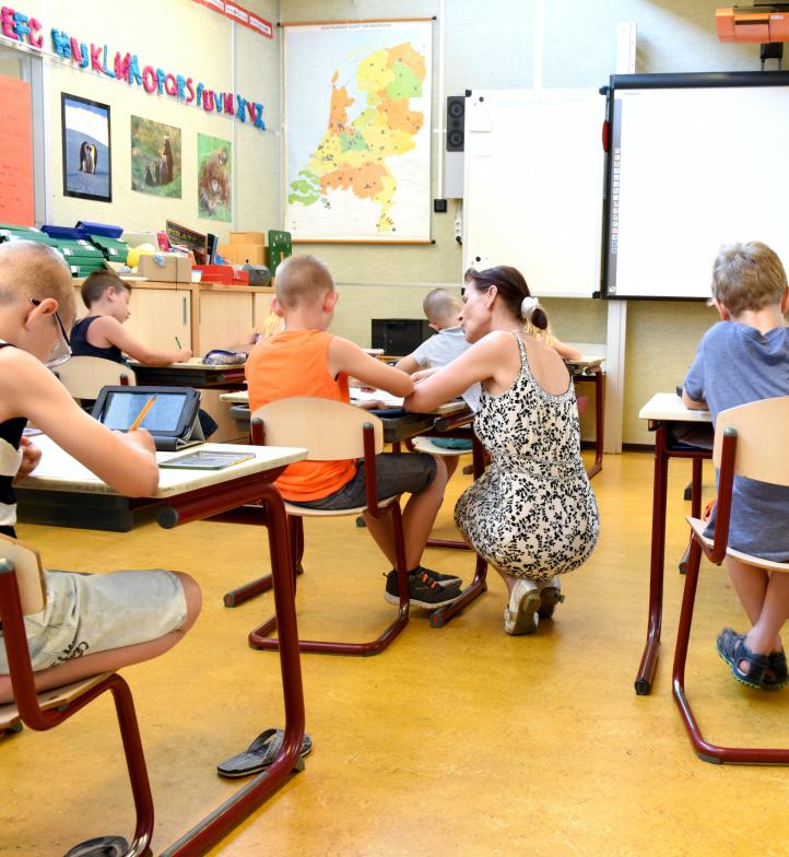 Schülerinnen und Schüler in einem Klassenzimmer. Eine Lehrerin unterstützt ein Mädchen.