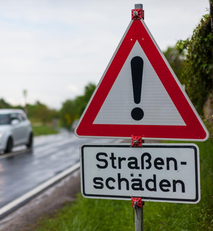 Bild einer kaputten Straße. Im Vordergrund steht ein Warnschild mit der Aufschrift "Straßenschäden"